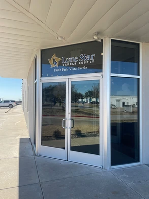 A glass double-door entrance to Lone Star Candle Supply, featuring the company logo and address "5800 Park Vista Circle" displayed above the doors. The entrance is part of a beige building with large windows, and a concrete sidewalk leads up to the doors. The reflection in the glass shows a parking lot and trees.