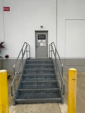 A white building exterior with a metal staircase leading up to a gray door labeled "Shipping & Receiving." The stairs have metal railings on both sides, and there are yellow bollards at the base of the stairs for protection.