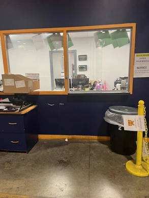 An interior office area with a navy-blue wall and a large glass window for customer interactions. A counter under the window holds paperwork and office supplies, while a trash can and caution sign stand nearby. A yellow post with a chain partially blocks access.