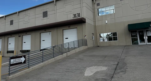 A beige industrial building with multiple roll-up doors numbered 13 through 16. A sloped ramp with a metal railing leads to a sign reading "Driver Entrance" with an arrow pointing toward the doors. The building has the number "3131" displayed near the top.