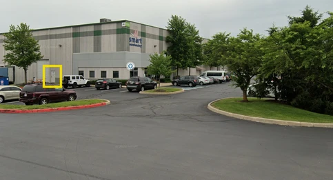 A parking lot in front of a large industrial building labeled "Smart Warehousing." Several cars are parked in the lot, and a door on the side of the building is highlighted with a yellow box.