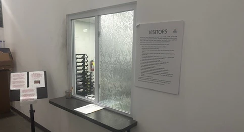 An interior area with a frosted glass window labeled for visitors. A sign on the wall outlines visitor policies, and a black counter below the window holds a clipboard and a cup with pens. A nearby stand displays additional notices.