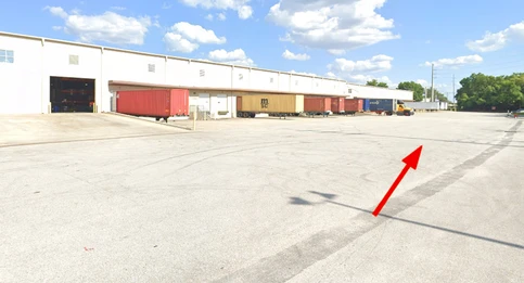A large truck court outside a white warehouse building with multiple loading docks and shipping containers parked along the docks. A yellow arrow on the pavement points toward the center of the lot, directing movement. The sky is clear with scattered clouds.