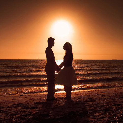 Couple silhouetted holding hands on beach at orange sunset — scent inspiration for Amber Romance fragrance oil