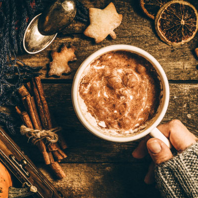 A cozy scene with a hand holding a warm mug of frothy hot chocolate or coffee, surrounded by autumn-themed items on a rustic wooden table. The setting includes cinnamon sticks, dried orange slices, star-shaped cookies, and a spoon, evoking a warm and inviting autumn atmosphere.