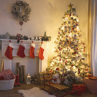 A cozy living room with a glowing Christmas tree beside a white fireplace hung with red stockings, surrounded by gifts, a teddy bear on a sled, and warm festive lights.
