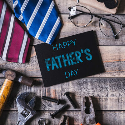 A festive Father's Day setup featuring a black greeting card with 'Happy Father's Day' written in teal letters, surrounded by an array of items such as a striped tie, glasses, and various hand tools like wrenches and pliers, all laid out on a rustic wooden surface.