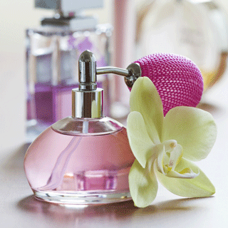 Glass perfume bottle filled with pink liquid, featuring a silver spray top and pink atomizer bulb, placed beside a pale yellow orchid flower.