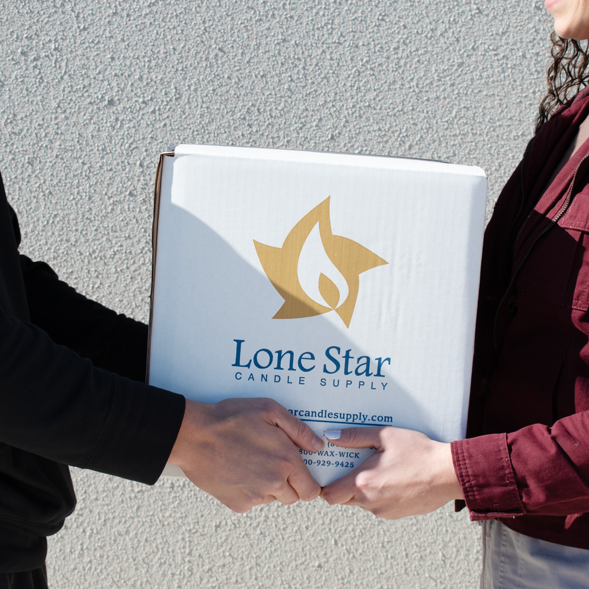 Two people exchanging a white box with the Lone Star Candle Supply logo, featuring a gold star and flame design, and the company's name in blue. The box also displays the website and contact information. One person wears a black jacket, and the other wears a burgundy jacket. The exchange takes place in front of a light gray textured wall.