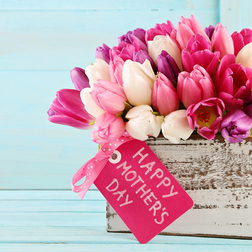 The image features a rustic white wooden container brimming with a vibrant assortment of tulips. The flowers, in shades of pink, fuchsia, and white, provide a beautiful contrast against the container's weathered texture. Attached to the blossoms is a festive pink tag with the words "Happy Mother's Day" inscribed in a cheerful, white font. The backdrop is a soft blue wooden surface, giving the composition a fresh and springtime feel.