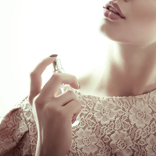 Close-up of a person’s lower face and neck as they hold a perfume bottle and spray fragrance, with soft lighting and a neutral background.