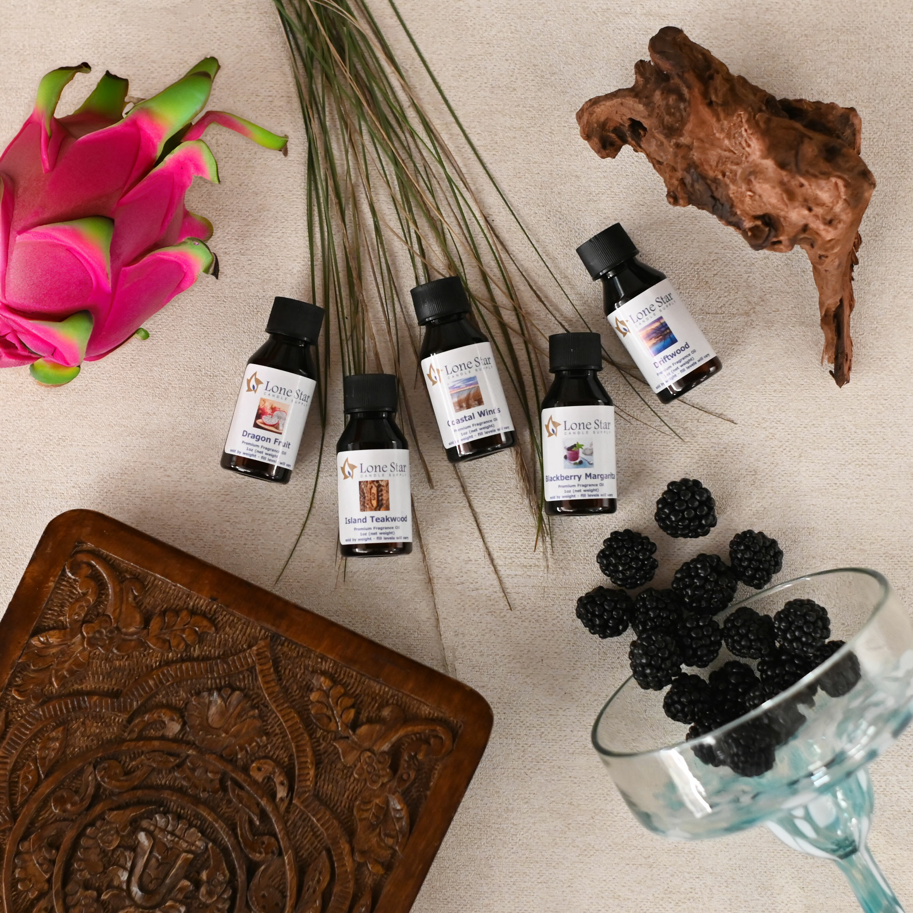 A flat lay image featuring bottles of Lone Star fragrance oils surrounded by a dragon fruit, blades of grass, a wooden carving, a piece of driftwood, and a glass filled with blackberries.