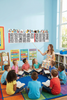 Sunday school teacher pointing to a colorful Books of the Bible wall decal while children sit in a circle reading Bibles in a bright Christian classroom.