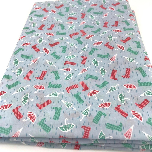 Image shows a sky-blue polycotton fabric printed with small red and green wellington boots and umbrellas. The pattern is evenly spaced across the fabric, which is 112cm wide with a smooth texture. Shown folded into a rectangle.