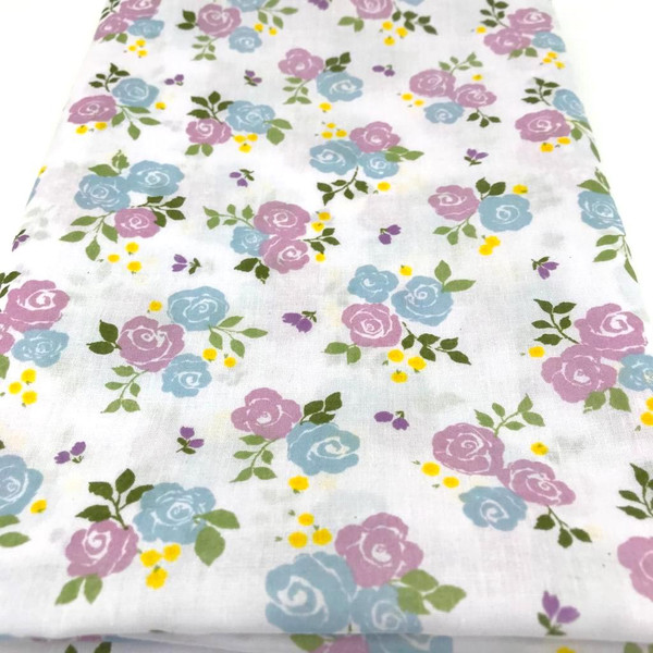 Image shows a white polycotton fabric printed with medium size lilac roses in clusters of three. The floral pattern is evenly spaced, creating a soft, summery look. The fabric is 112cm wide with a smooth finish and shown folded in a rectangle.