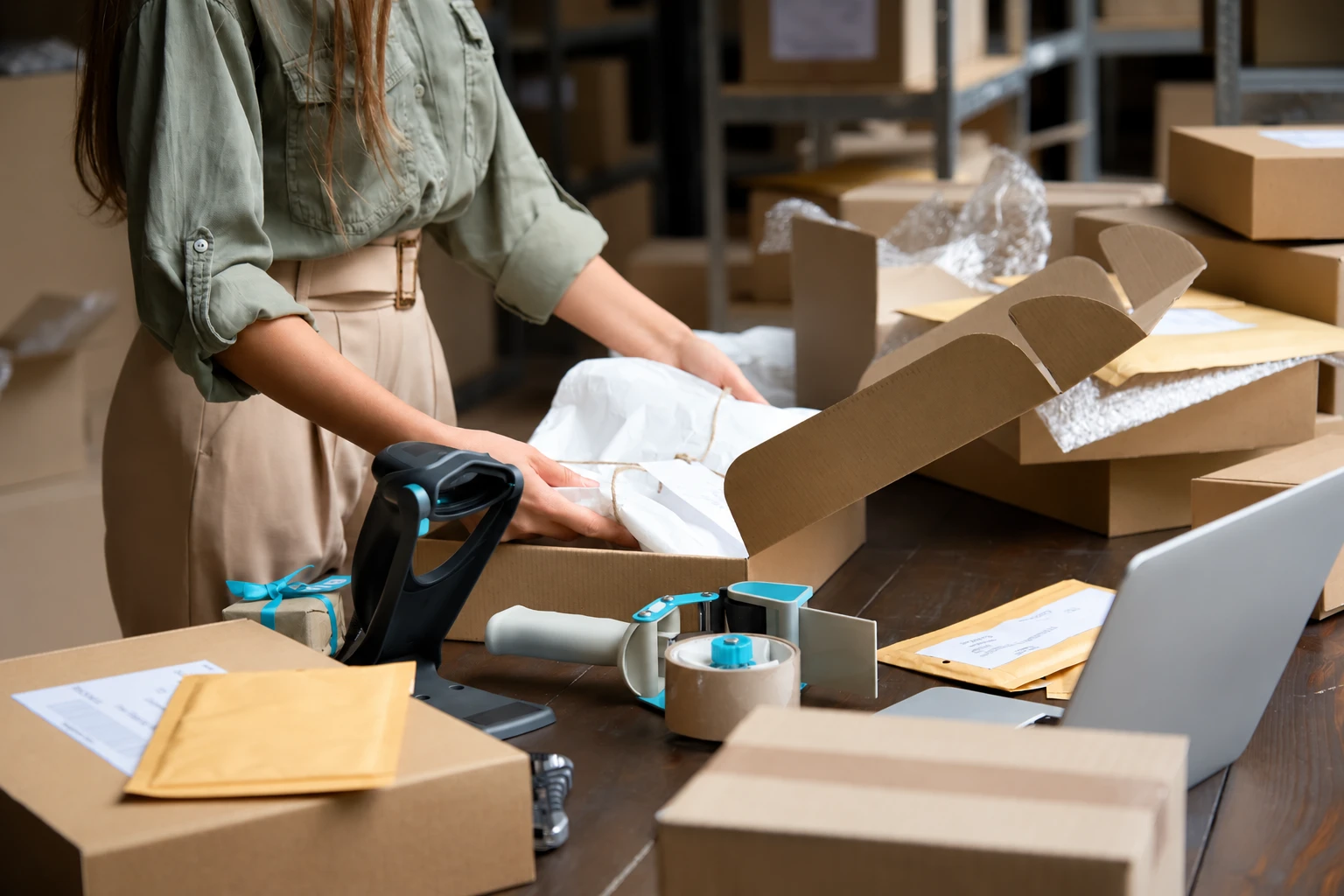 Woman packing a tissue-wrapped item into a cardboard shipping box at a workstation with mailers, packing tape, and a laptop.