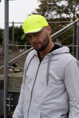 A man wearing a bright yellow cap stands outdoors, paired with a grey hoodie. The cap features reflective trim and a logo.