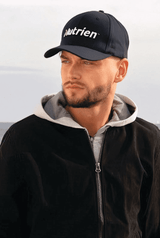 A navy cap with a logo, worn by a person in a grey hoodie and black jacket, set against a natural background.