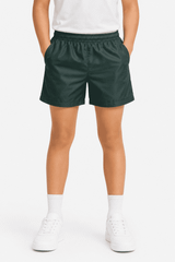 Kids' sports shorts in dark green with an elastic waistband and side pockets, paired with white shoes and socks.
