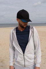 A man wearing a black cap with a blue peak, grey zip-up hoodie, and navy t-shirt stands on a beach.