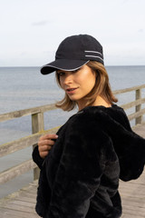 A black microfibre sports cap with white piping, worn by a woman by the seaside. The setting includes a wooden railing.