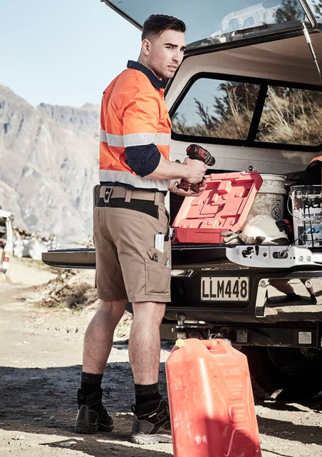 A man wearing orange and navy workwear shorts stands beside a vehicle, holding a drill and a red toolbox.