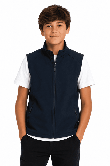 A boy stands wearing a navy blue vest over a white T-shirt, with his hands in his pockets. The vest has branding.
