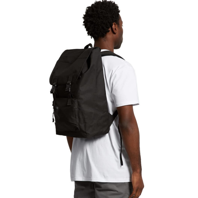 A black recycled field backpack worn over a person's shoulder, featuring adjustable straps and a simple design.
