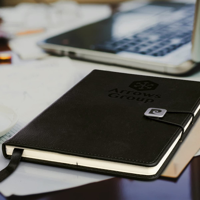 A black notebook with a logo and a clasp, placed on a desk beside a laptop and scattered documents.