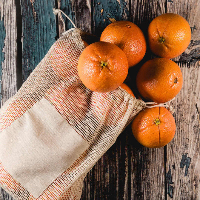 A reusable mesh produce bag containing several bright orange oranges, resting on a wooden surface.