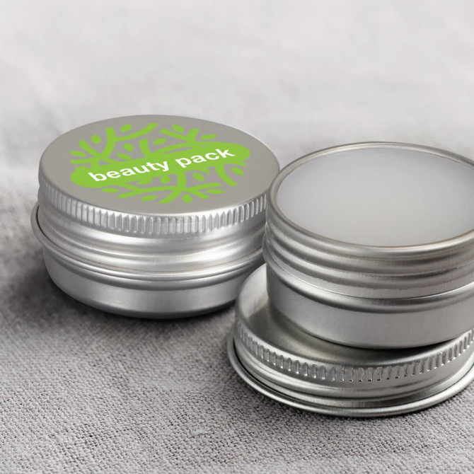 A silver tin for lip balm with a green decorative lid, featuring a logo. Two tins are partially open on a grey fabric background.