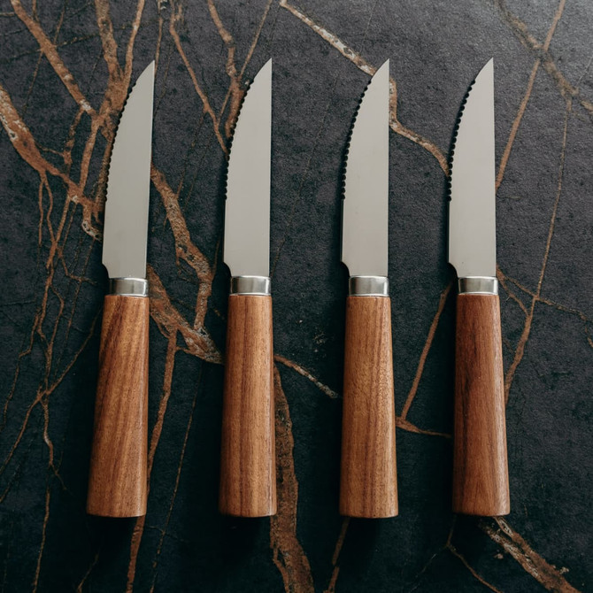 A steak knife set featuring four stainless steel knives with brown wooden handles, arranged on a dark stone surface.
