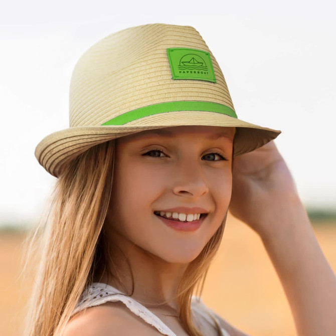 A straw fedora hat in beige with a green band and logo, worn by a smiling young girl outdoors.