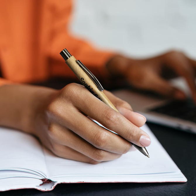 A plastic pen with a brown barrel and silver accents is held above a notebook. The background features a laptop.