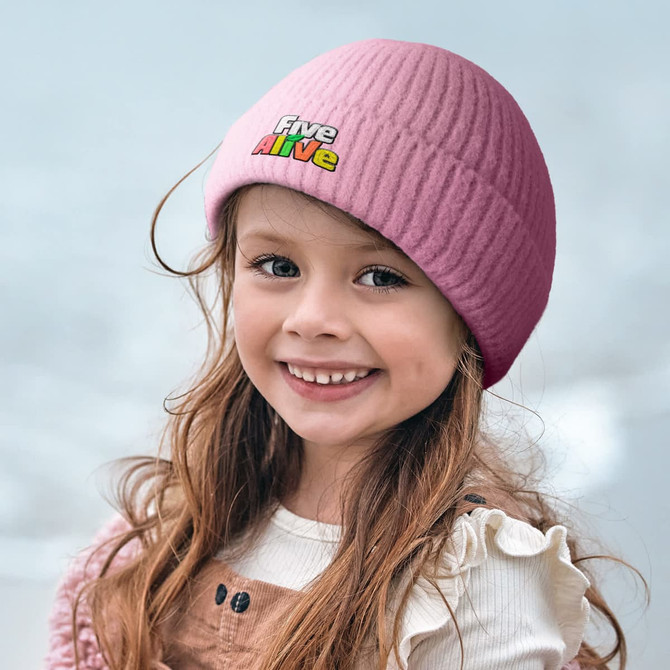 A pink brushed kids beanie sits atop a smiling girl's head, featuring a logo on the front.