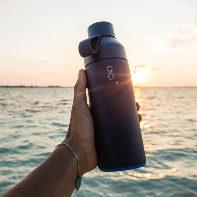 A drink bottle in dark blue with a lid, held against a sunset over a body of water, featuring a logo.