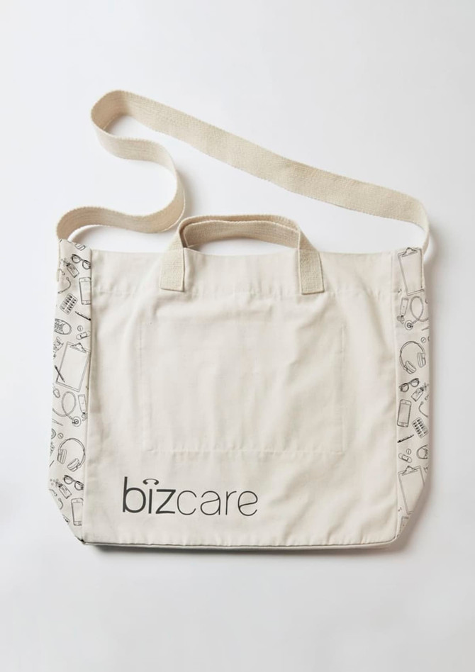 A beige unisex tote bag featuring a patterned edge and a logo. It has two carrying handles and a shoulder strap.