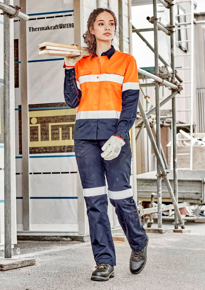 A woman wearing orange and navy workwear, featuring a utility shirt and pants, stands on a construction site holding timber.