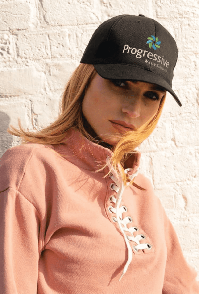 A black organic brushed heavy cotton cap with a logo, worn by a model in a pink pullover against a white wall.