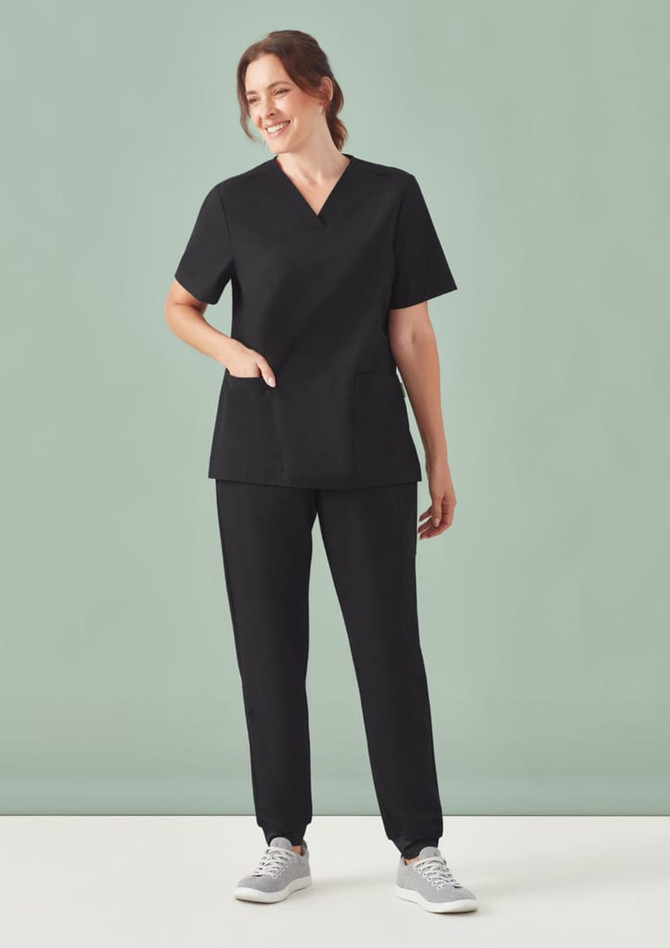 A woman wearing a black V-neck scrub top and matching pants, with a pocket, set against a green background.