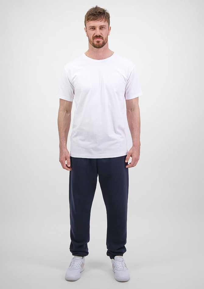 A man stands wearing a white t-shirt and navy blue track pants. The pants have a logo on them.