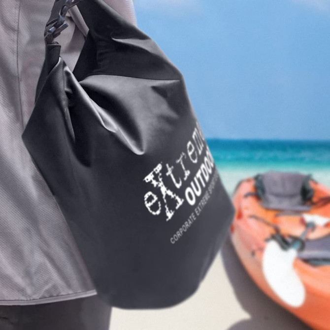 A 10L black dry bag with a logo, held by a person near a sandy beach and kayak in the background.