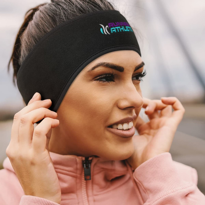 A black ear warmer is being worn by a woman, featuring a logo on the front. She is adjusting it with her fingers.