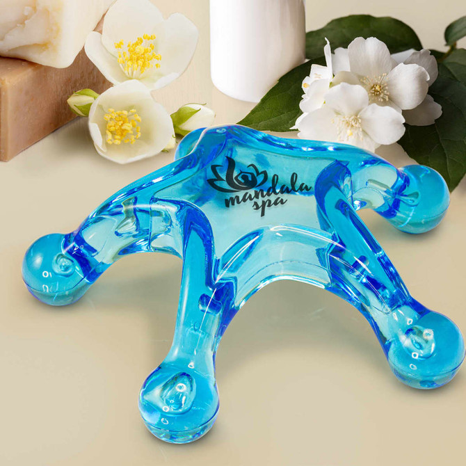 A blue star-shaped massager with rounded ends, placed near white flowers and bars of soap. It has a logo.