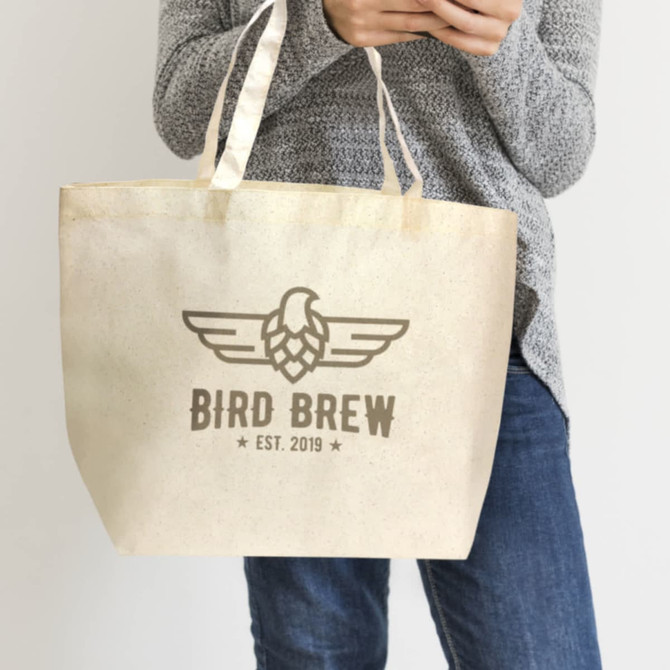 A natural-coloured tote bag with a logo, held by a person wearing a grey sweater and jeans.
