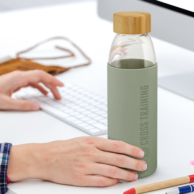 A drink bottle with a glass body, green silicone sleeve, and bamboo lid, resting on a desk next to a computer.