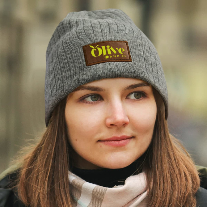 A grey cable knit beanie featuring a patch. The beanie is worn by a person with long hair.