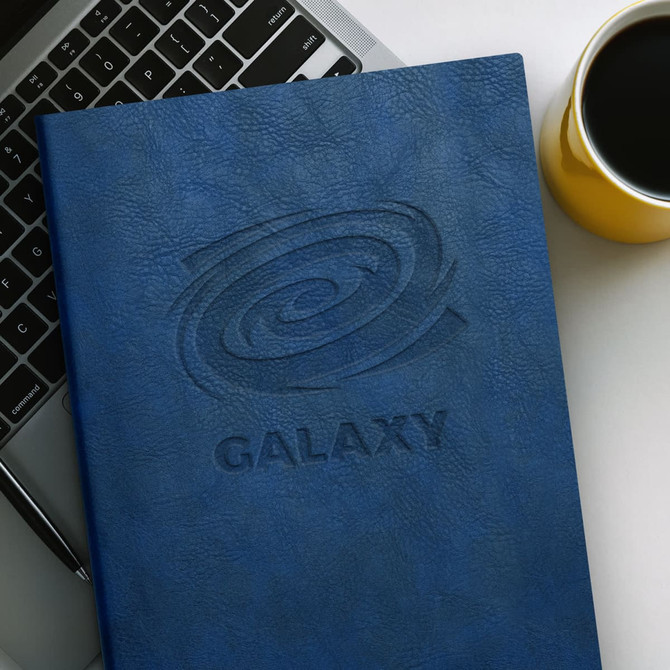 A large blue soft cover notebook with an embossed logo, placed near a laptop and a yellow coffee cup.