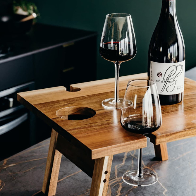A wooden folding wine table featuring two glasses and a bottle of red wine, set against a dark green background.