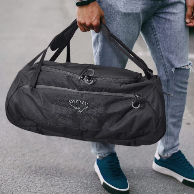 A black duffle bag with a logo, featuring dual handles, is being held by a person in casual attire.